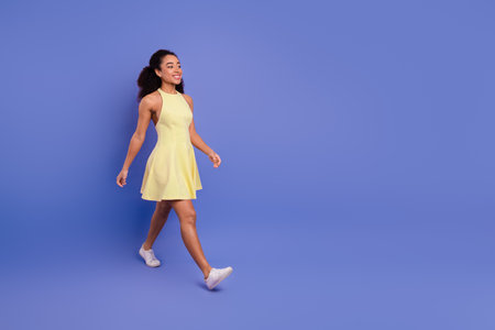 Energetic young woman in a yellow dress confidently walking with a smile against a vibrant purple backgroundの写真素材