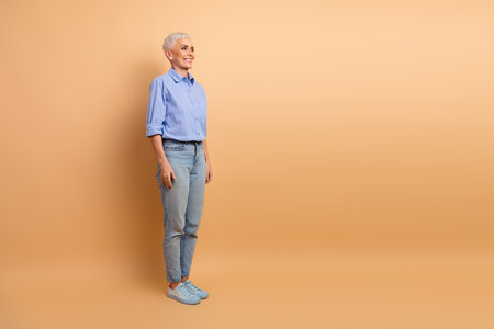 Confident mature woman posing in casual attire with a beige background, embracing elegance and personal style with a cheerful demeanorの写真素材