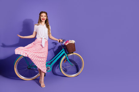 Young female enjoying leisure time near bicycle with flowers on purple background in casual and stylish outfit, smiling.の写真素材