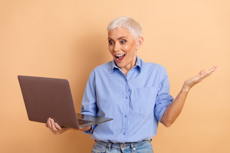 Excited Mature Woman Using a Laptop for Online Business on a Beige Background
