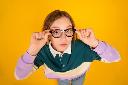 Focused young woman in trendy casual clothing adjusting glasses against a bright yellow backgroundの写真素材