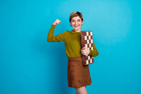 Confident young woman holding chessboard in retro casual outfit against bright blue background...
