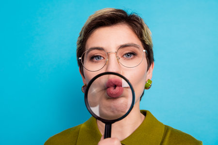 Young businesswoman holding magnifying glass over her lips against bright blue background showcasing her personalityの写真素材