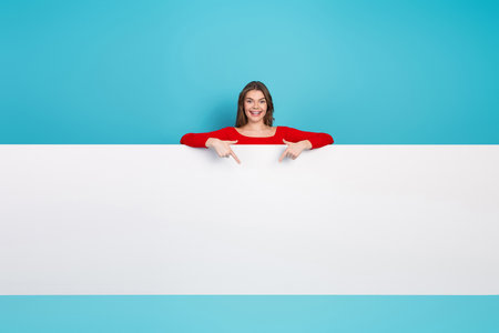 Young cheerful woman pointing down against a blue background showcasing a blank white panel for text or advertisingの写真素材