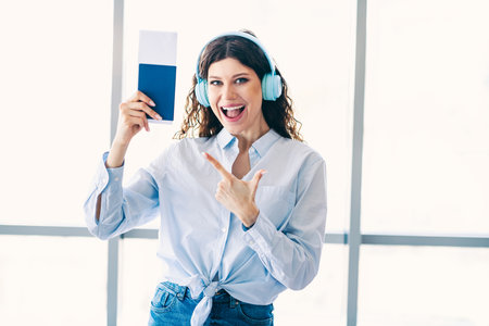 Excited young woman in casual attire holding travel ticket with headphones ready for a new journey and destination adventureの写真素材