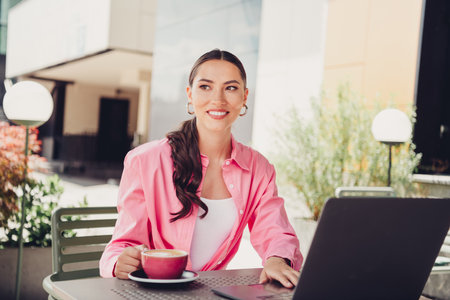 Lovely woman enjoying leisure time outdoors with laptop and coffee exploring cityscape in a casual and stylish outfitの写真素材