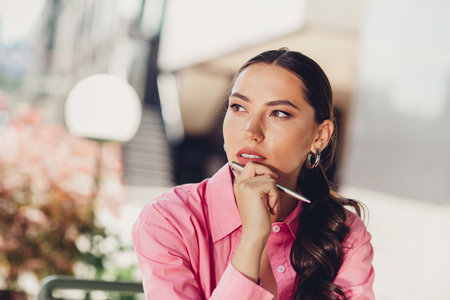 Charming woman in pink shirt outdoors during daylight, pondering thoughtfully while holding a pen, surrounded by urban sceneryの写真素材