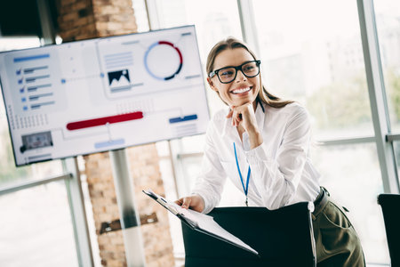 Charming young businesswoman analyzes data on a digital screen in a modern office with professional attireの写真素材