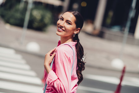 Urban scene featuring a stylish young woman walking outdoors on a sunny day, wearing a pink shirt and smiling brightlyの写真素材