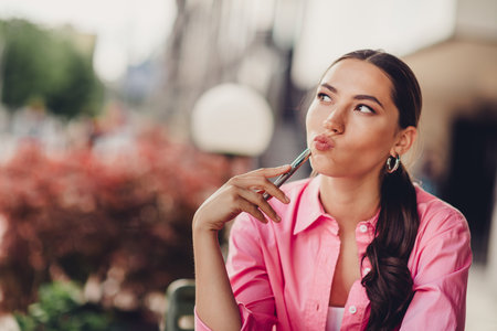 Charming woman in a pink shirt reflecting outdoors in a city setting, holding a pen, during a sunny, relaxing day in autumnの写真素材