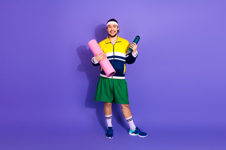 Young cheerful man in retro sportswear holding a yoga mat and water bottle against a vibrant purple backgroundの写真素材