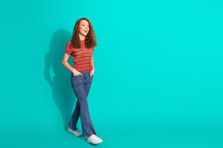 Smiling young woman in stylish casual attire posing against a vibrant turquoise background with hands in pocketsの写真素材