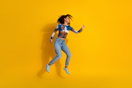 Energetic young woman jumps joyfully against a vibrant yellow background showcasing casual style and...