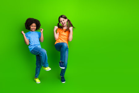 Two happy preteen girls celebrating and dancing in casual outfits against a vibrant green backgroundの写真素材