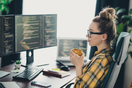 Female programmer working on coding tasks at a modern workspace with multiple computer screens and...