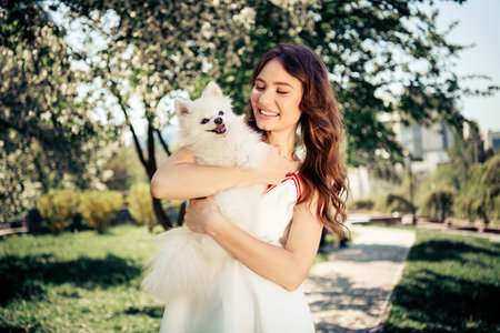 Young woman holding a fluffy white dog in a blooming park during a sunny spring day, enjoying an outdoor leisure walk.の写真素材