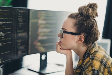 Focused young female programmer working remotely on a dual-screen setup in a casual home office environmentの写真素材