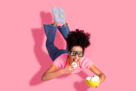 Cheerful young woman with curly hair enjoying snacks while laying on vibrant pink background.