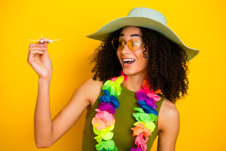 Smiling woman in summer hat holding toy airplane against a bright yellow background, celebrating joy and travelの写真素材