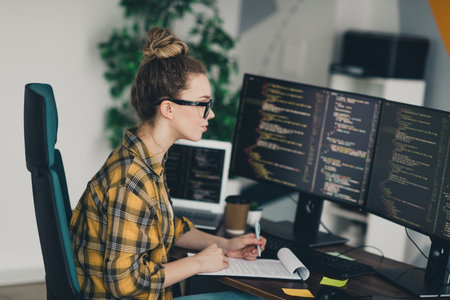 Confident female programmer working on software code using multiple screens in a modern office workspaceの写真素材