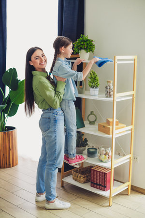 Mother and daughter cleaning bookshelf together in cozy home environment during a leisure day, creating bonding momentsの写真素材