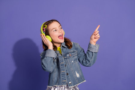 Happy little girl wearing headphones and denim jacket enjoying music and dancing on a purple...