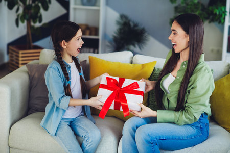 Mother and daughter sharing a joyous moment while exchanging a gift on their cozy homes sofa.の写真素材