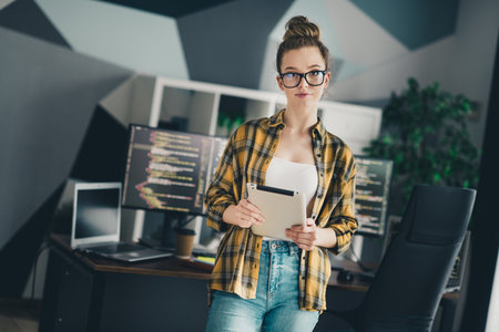 Confident female programmer holding a tablet in a modern workspace surrounded by computer screens...