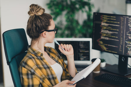 Young female programmer focusing on a coding project in a modern office workspace with multiple...