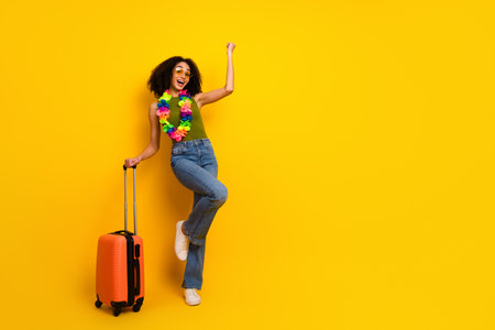 Joyful young woman with colorful lei and orange suitcase enjoying holiday vibes on a bright yellow backgroundの写真素材