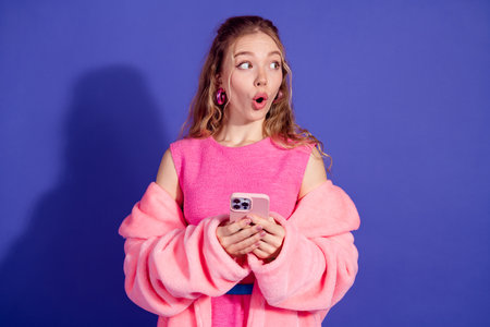 Young woman wearing a pink outfit holding a phone with surprise expression against purple backgroundの写真素材