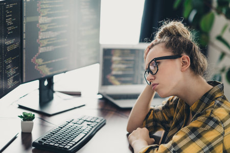 Female programmer analyzing code on desktop monitor while working remotely in a home office setting on software development tasksの写真素材