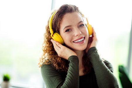 Charming young woman enjoying music with headphones in a comfortable indoor setting, showcasing casual style and happinessの写真素材
