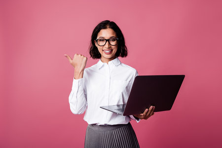Young businesswoman in formal white shirt holding a laptop with a cheerful expression against pink backgroundの写真素材