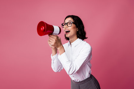 Joyful businesswoman with megaphone making announcement dressed in formal attire against vibrant pink backgroundの写真素材