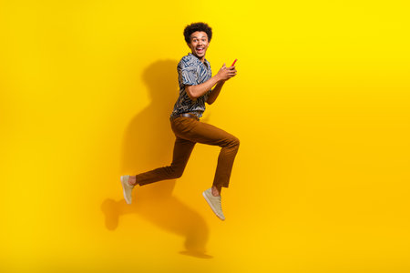 Excited young man in stylish outfit happily leaping with phone against vibrant yellow background, expressing joy and energyの写真素材