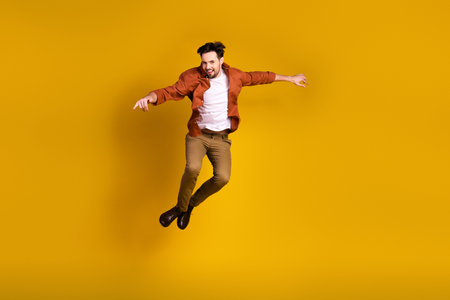 Dynamic portrait of a joyful young man in casual attire jumping energetically on a vibrant yellow backdropの写真素材