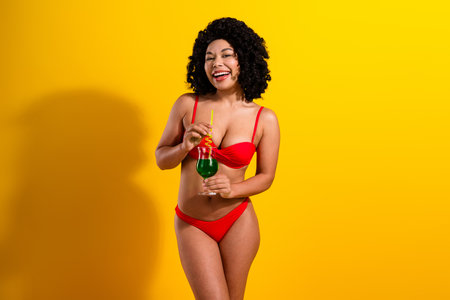 Young woman in vibrant red swimwear enjoying a fun moment with a cocktail on a bright yellow backgroundの写真素材
