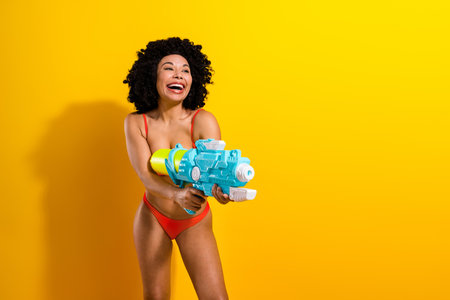 Cheerful woman in red swimsuit enjoying summer fun holding a colorful water gun against a bright yellow backgroundの写真素材