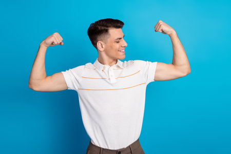 Confident young man showing off his strength in a stylish white shirt against a vibrant blue...