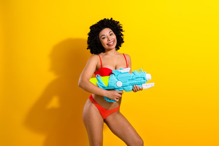 Smiling young woman in red bikini holding large water gun against yellow background expressing fun and vibrant summer vibesの写真素材