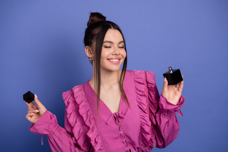 Beautiful young woman in pink ruffled dress holding perfume bottle on purple background promoting...