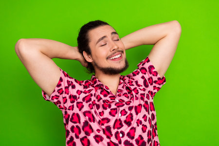 Confident young man in trendy leopard print shirt posing with hands on head against vibrant green...