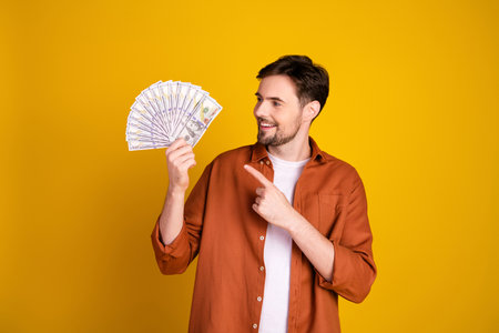 Young man in casual orange shirt on yellow background holding money and pointing, expressing wealth and successの写真素材
