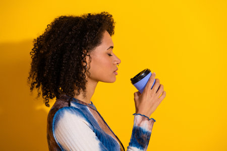 Stylish woman with curly hair enjoying a moment while holding a coffee cup against a bright yellow...