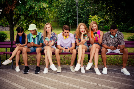 A diverse group of young friends sitting together on a park bench enjoying a sunny day and bonding...