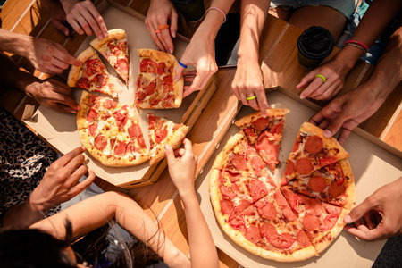 Group of friends sharing pizza outdoors on a sunny day, celebrating togetherness and enjoying leisure momentsの写真素材
