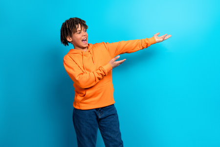 Cheerful boy in a bright orange hoodie gesturing against a vibrant blue background, expressing happiness and energyの写真素材