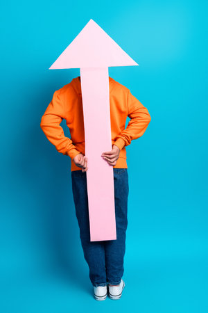 Young boy in orange hoodie holding upward pink arrow on blue background representing progress and growthの写真素材