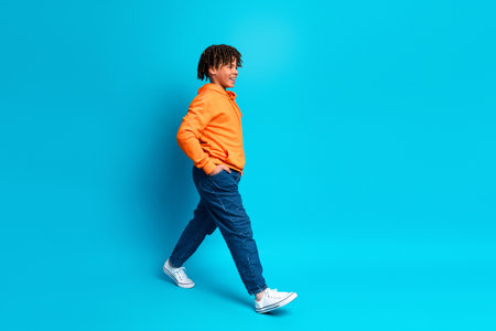 Smiling young schoolboy in a vibrant orange hoodie strolling confidently against a blue studio background, full-body profile shotの写真素材
